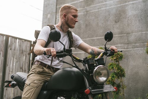 Free A stylish, tattooed young man confidently sits on a vintage motorcycle against an outdoor urban background. Stock Photo
