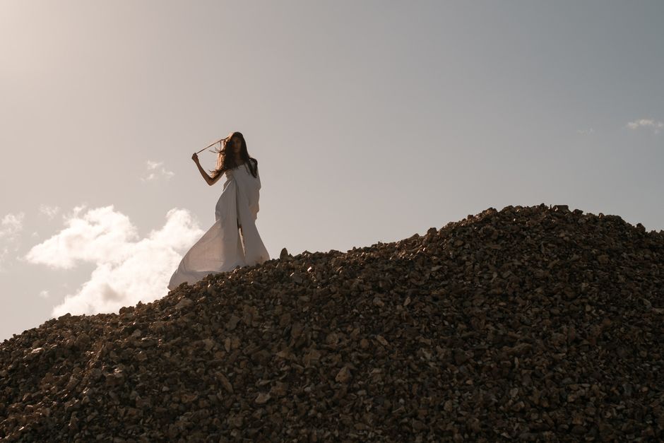 From below of unrecognizable female wearing white long dress touching hair while standing on edge of hill