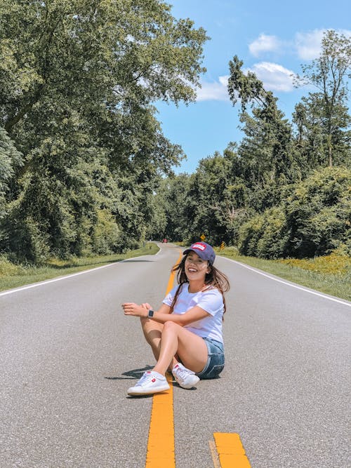 Full length of smiling young female enjoying summertime while sitting on asphalt road and laughing