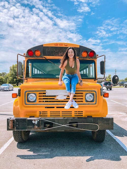 Full body of happy young female sitting on hood of colorful school bus in parking lot