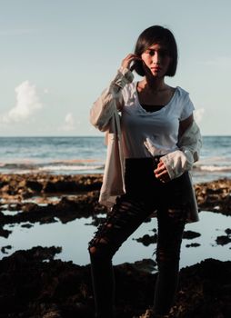 Young ethnic female in casual clothes touching hair while standing on stony coast against blue sea