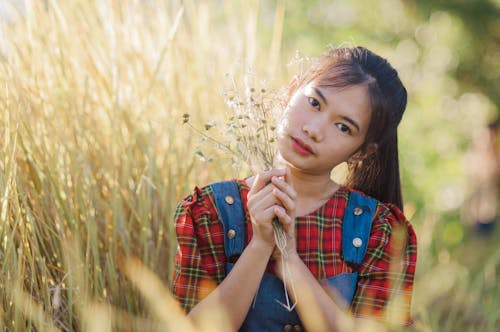 Calm Asian adolescent with bunch of flowers in long meadow grass looking at camera