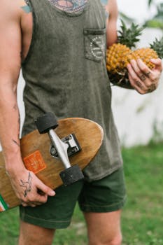 Crop anonymous fit man in tank top and shorts with skateboard and pineapples in hands standing against blurred backdrop of backyard in summer