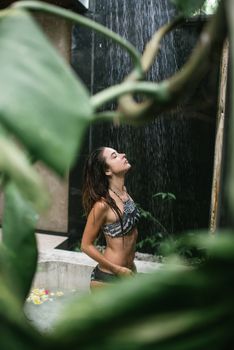 Side view of slender lady in bathing suit enjoying warm shower spray while standing in resort pool with exotic greenery