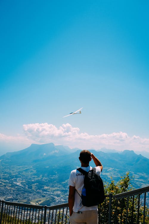 Back view of anonymous backpacker standing on observation deck and enjoying spectacular mountain range and aircraft flying in blue sky