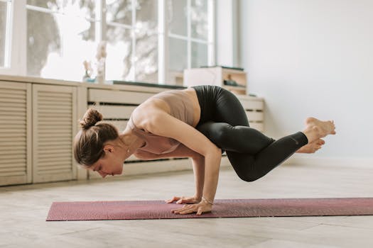 Woman holds an arm balance yoga pose on a mat in a bright room.