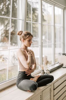 Young woman practicing meditation in a serene yoga pose by a sunlit window indoors.