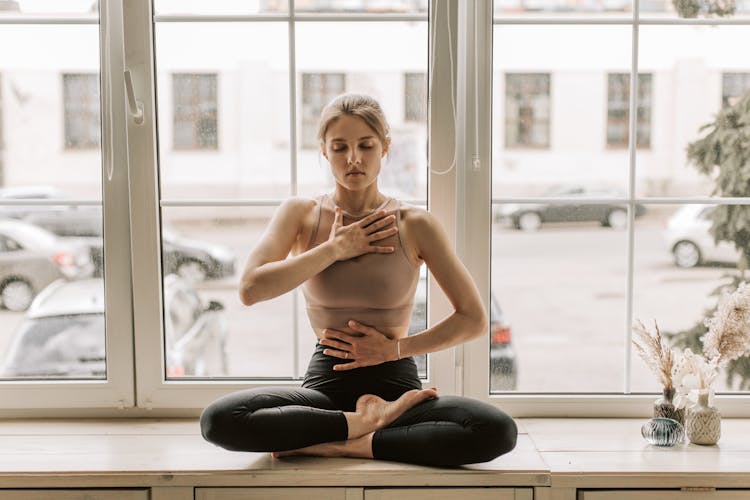 A Woman In Black Leggings Sitting Near The Glass Windows While Meditating