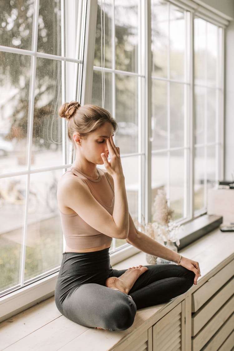 A Woman In Black Leggings Sitting Near The Window While Meditating