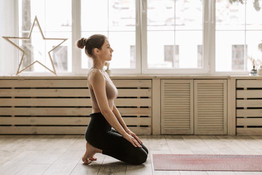 Woman in a yoga pose, kneeling on a wooden floor in front of large windows, embracing wellness.