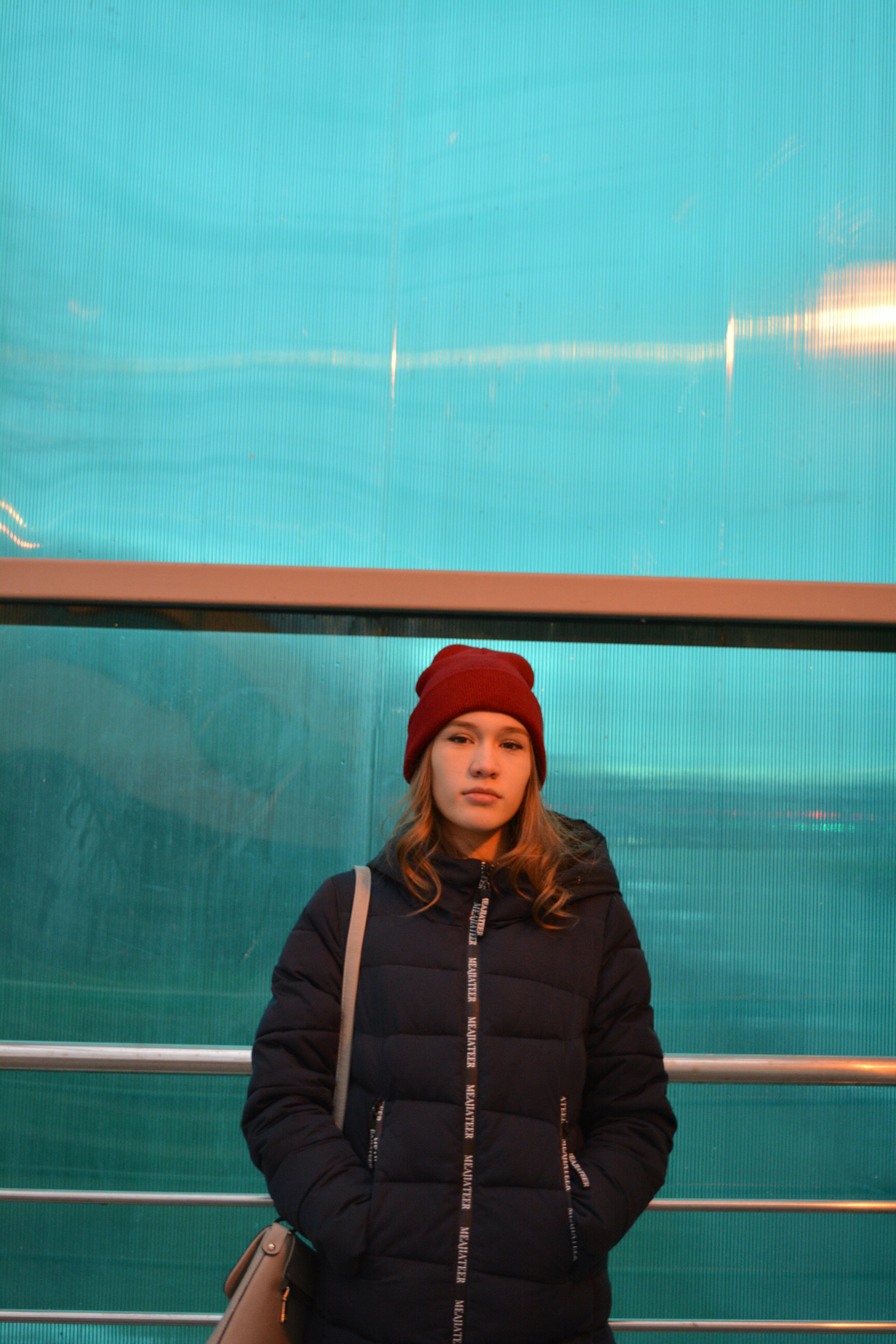 Serious young female in warm winter black coat and red hat looking at camera while standing by fence next to blue wall with hands at pockets in light of lamps