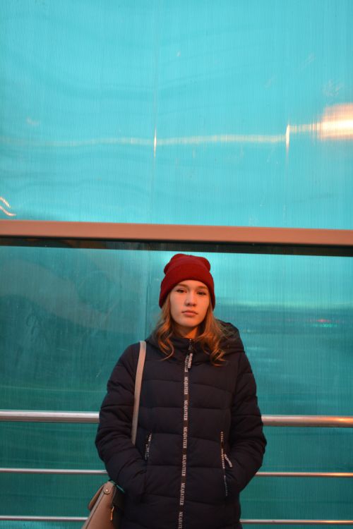 Free Serious young female in warm winter black coat and red hat looking at camera while standing by fence next to blue wall with hands at pockets in light of lamps Stock Photo