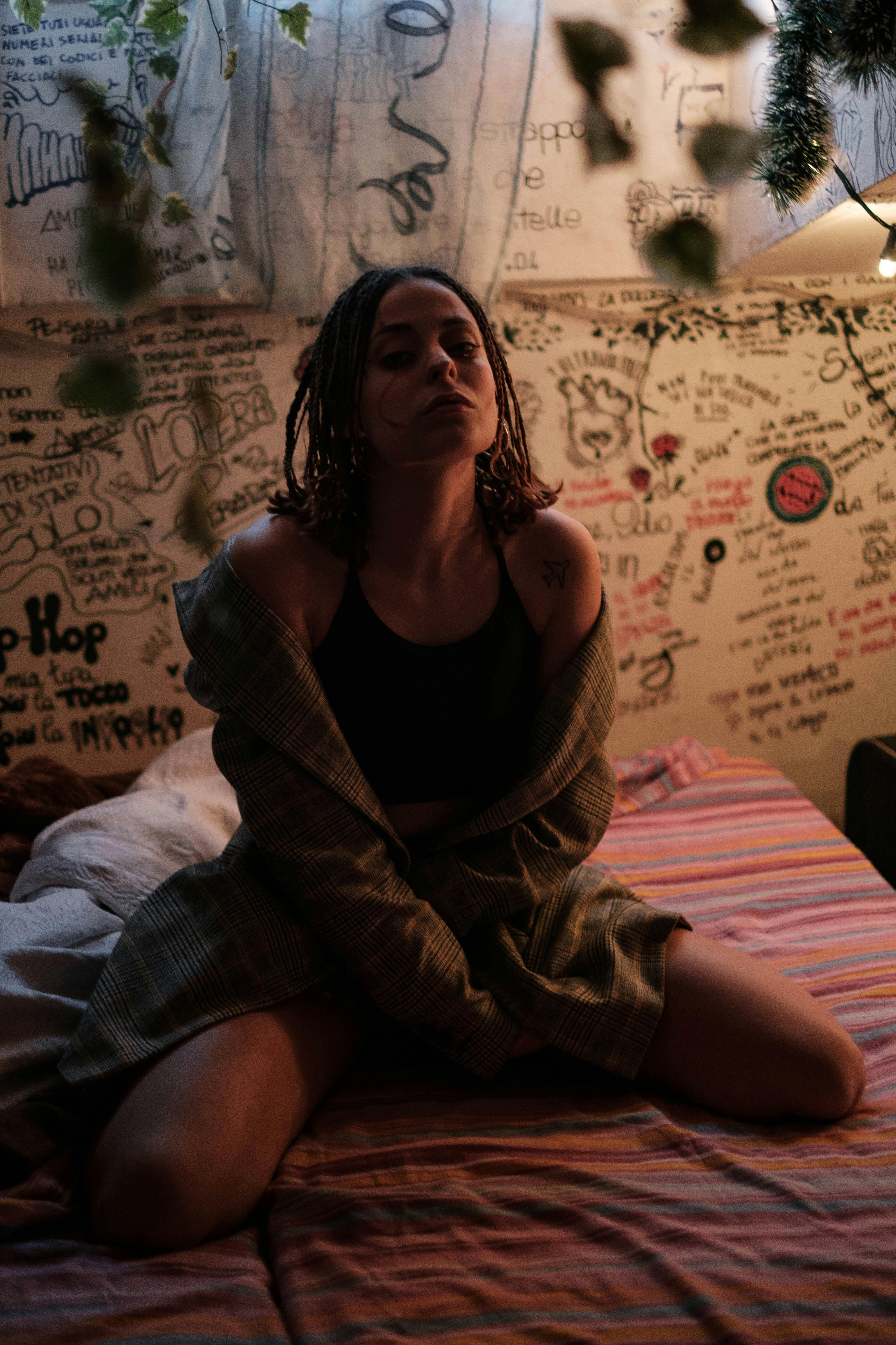 From above of young sensitive ethnic lady sitting on creased bed sheet near decorative wall at night at home