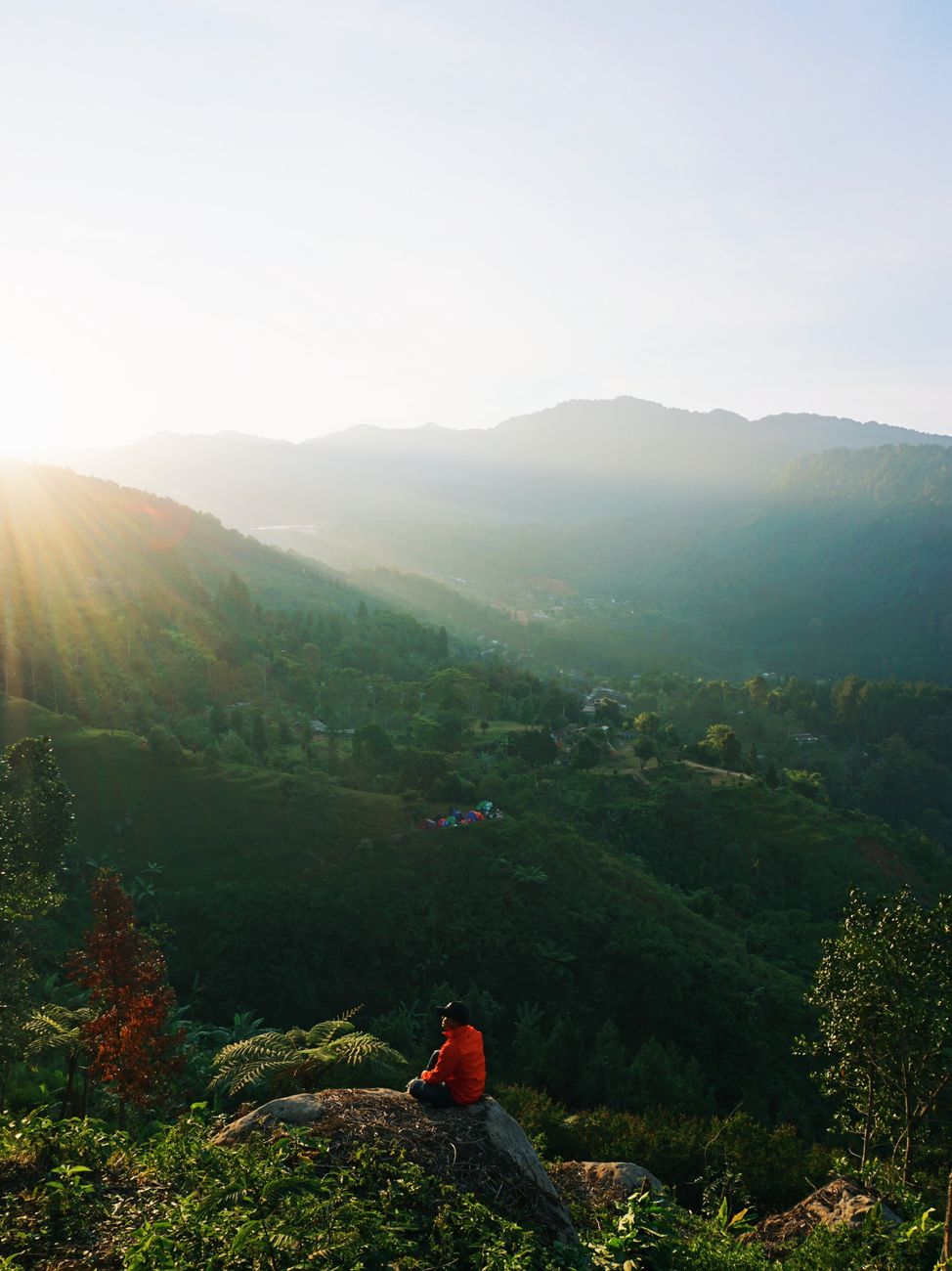 From above back view of anonymous tourist on stone admiring green ridge under cloudy sky at sunrise