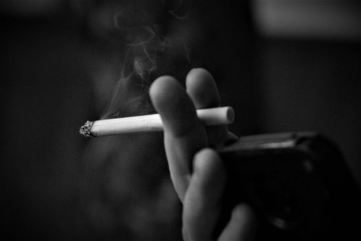 黑白照片中，一只手夹着香烟，烟雾缭绕，背景模糊