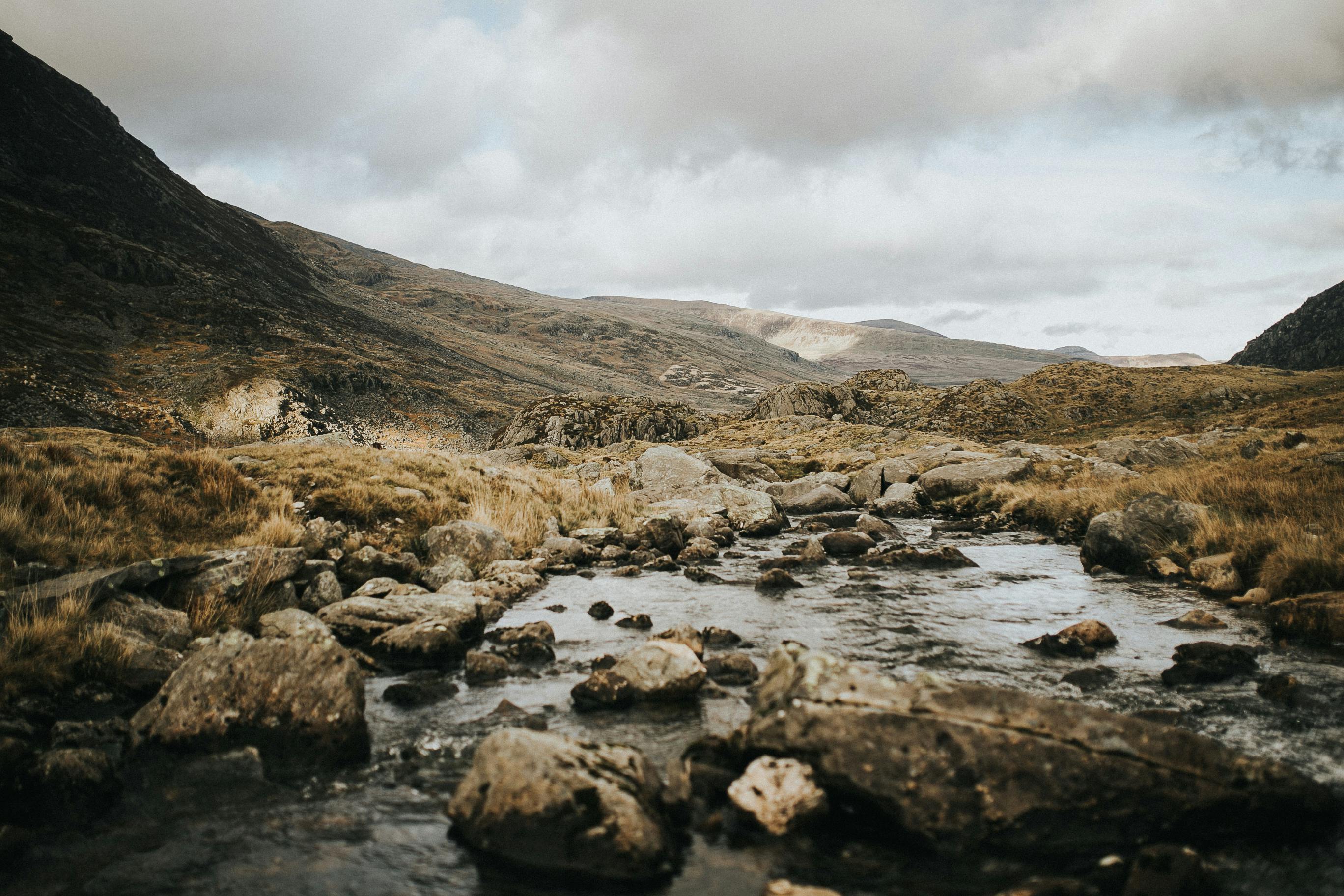 Serene mountain landscape with a gentle stream and rocky terrain under cloudy skies.
