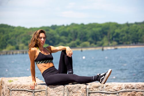 Free Side view of slim sportswoman in active wear with abdomen sitting on cement fence near ocean while looking at camera after working out Stock Photo