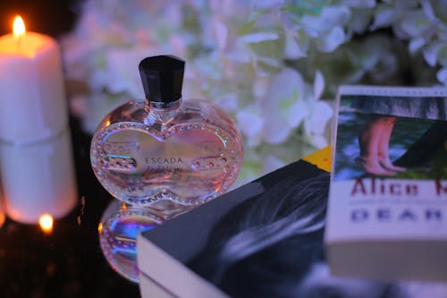 From above of ornamental bottle of aromatic perfume near bright candle and books behind blooming flowers at home