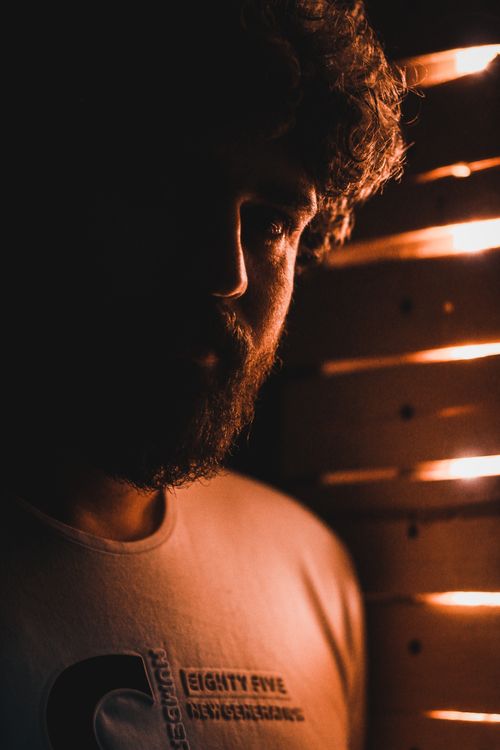 Free Crop unshaven male with curly hair in casual wear standing near window at home in sunlight while looking at camera Stock Photo