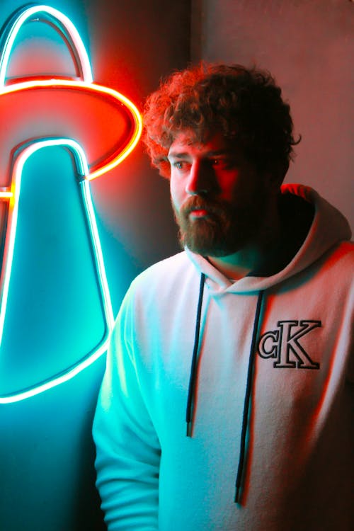 Bearded Man with Curly Hair in White Hoodie Standing Beside Neon Sign