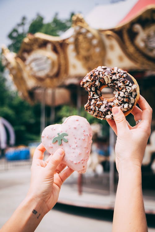Person Holding Chocolate Doughnut With Nuts and Heart Shaped Cookie Beside Carousel 