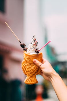 A fish-shaped waffle cone with ice cream held by a hand outdoors, showcasing street food delight.