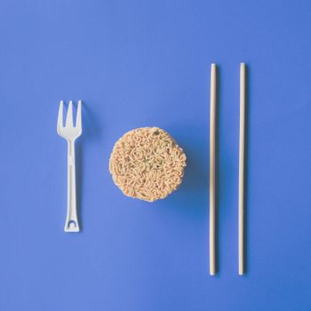 Top view composition of delicious noodles and plastic fork with wooden chopsticks arranged on blue background