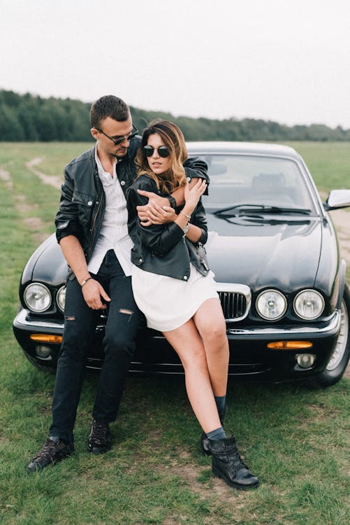 Full body stylish man and woman in leather jackets embracing near old fashioned automobile in countryside