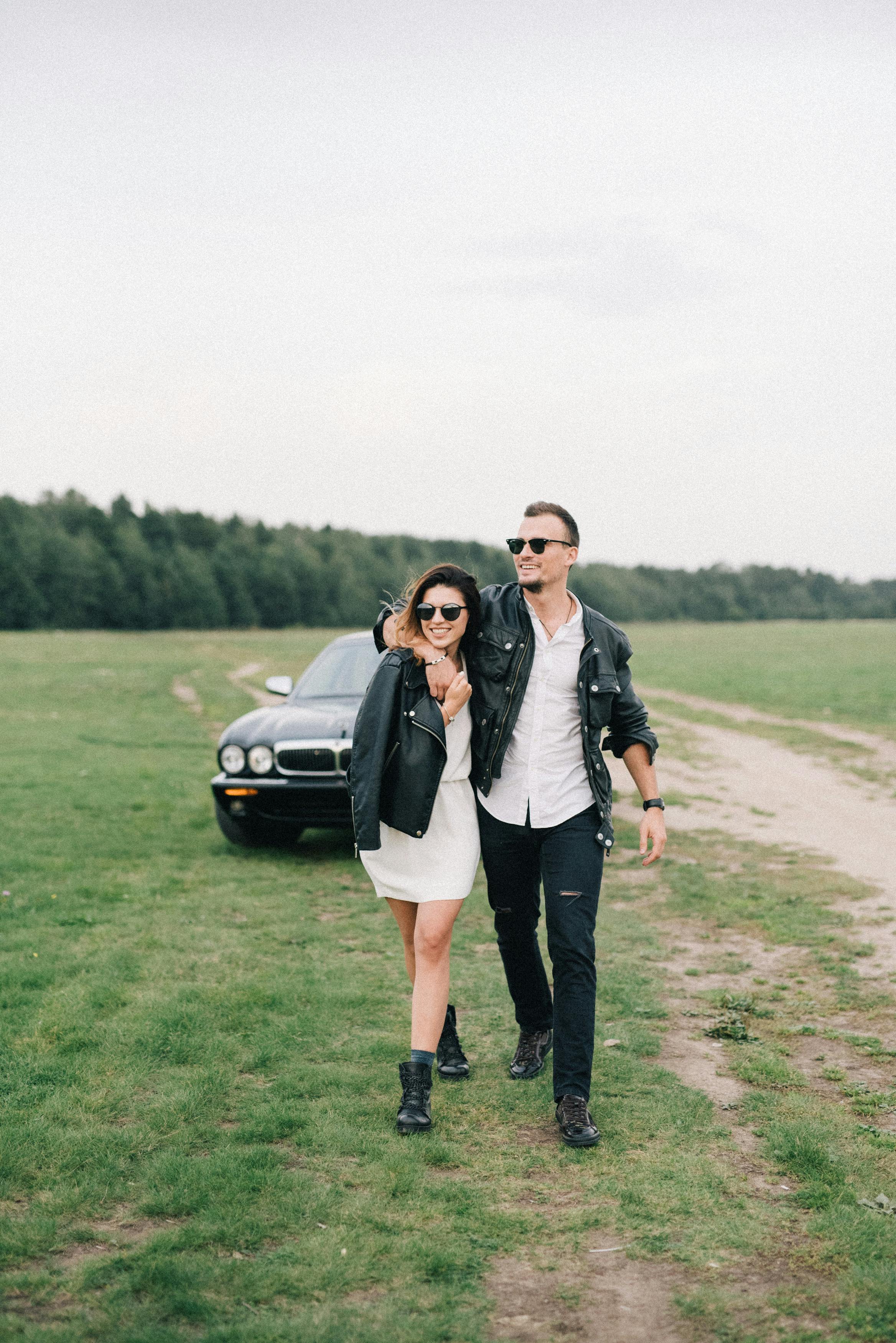 一对时尚情侣穿着黑色皮夹克走过草地，背景是一辆汽车