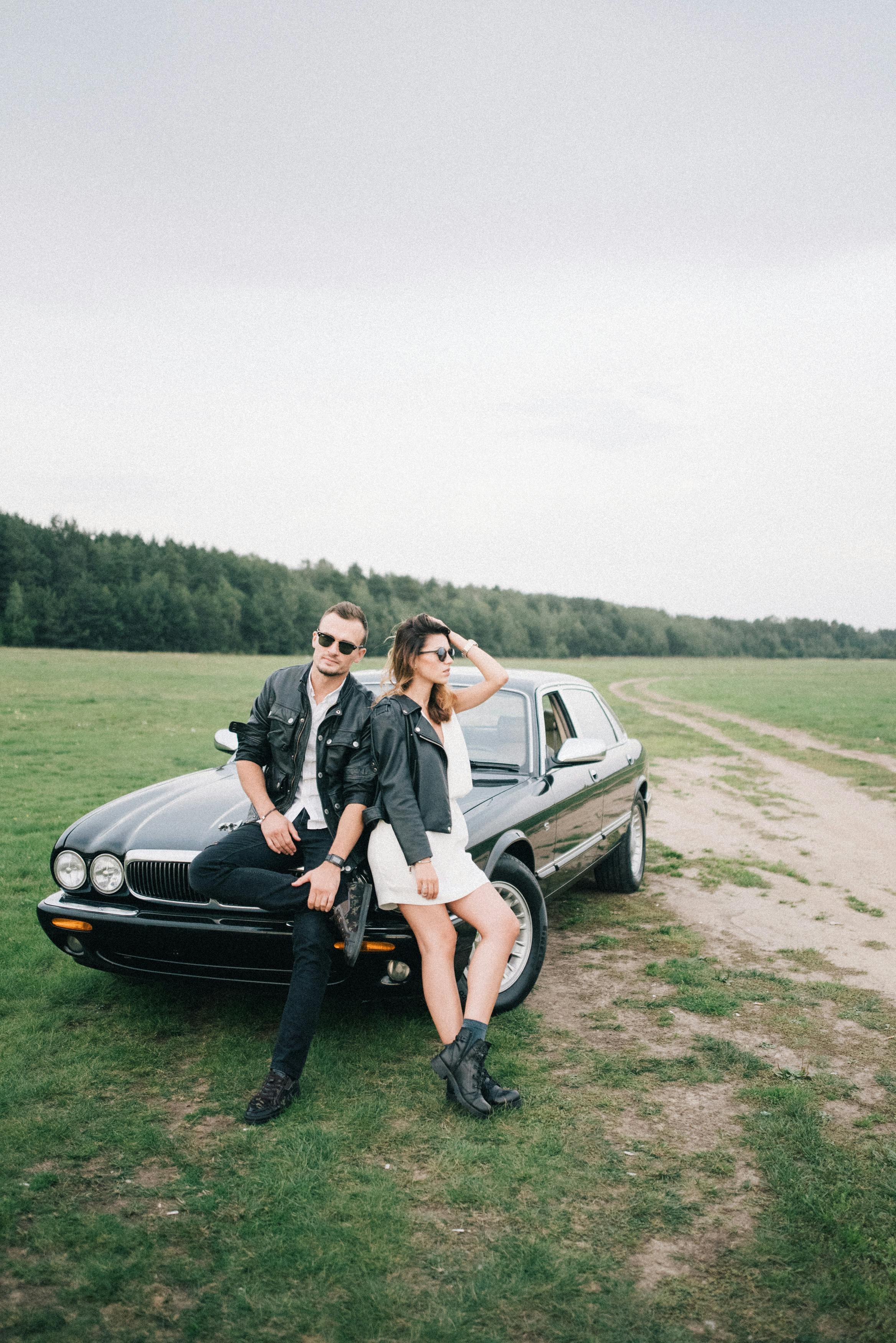 一对穿着皮夹克的时尚情侣，在一望无际的田野上与一辆老式黑色汽车合影