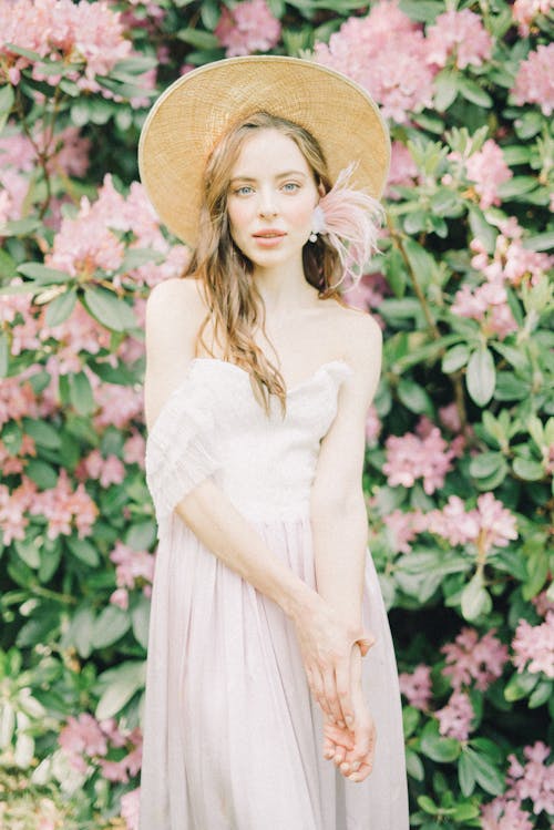 Femme En Robe Blanche Sans Manches Portant Un Chapeau De Soleil Marron Debout Près De Fleurs Roses