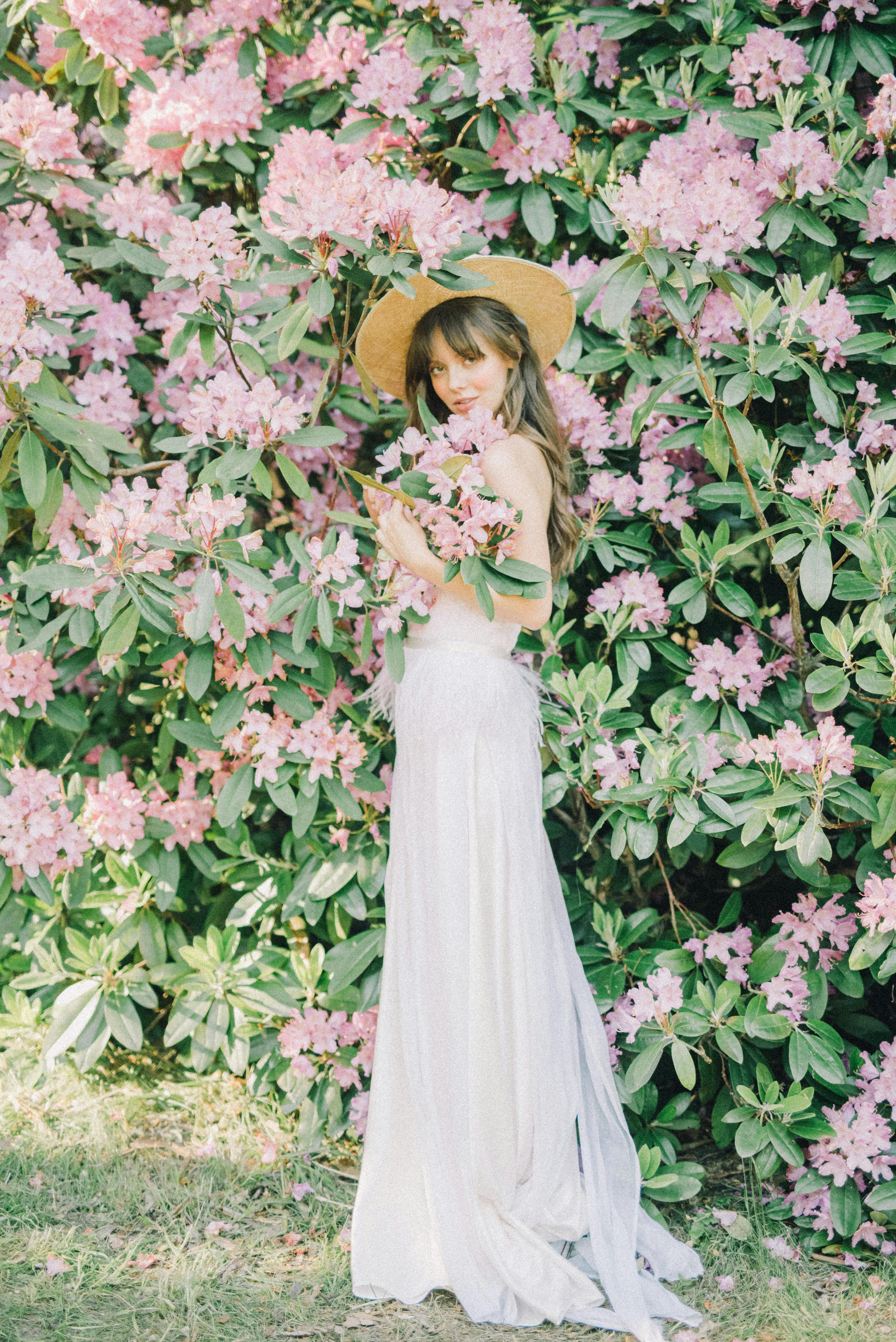 一位身穿白色连衣裙、头戴遮阳帽的年轻女子，正在花园里与粉色花朵合影