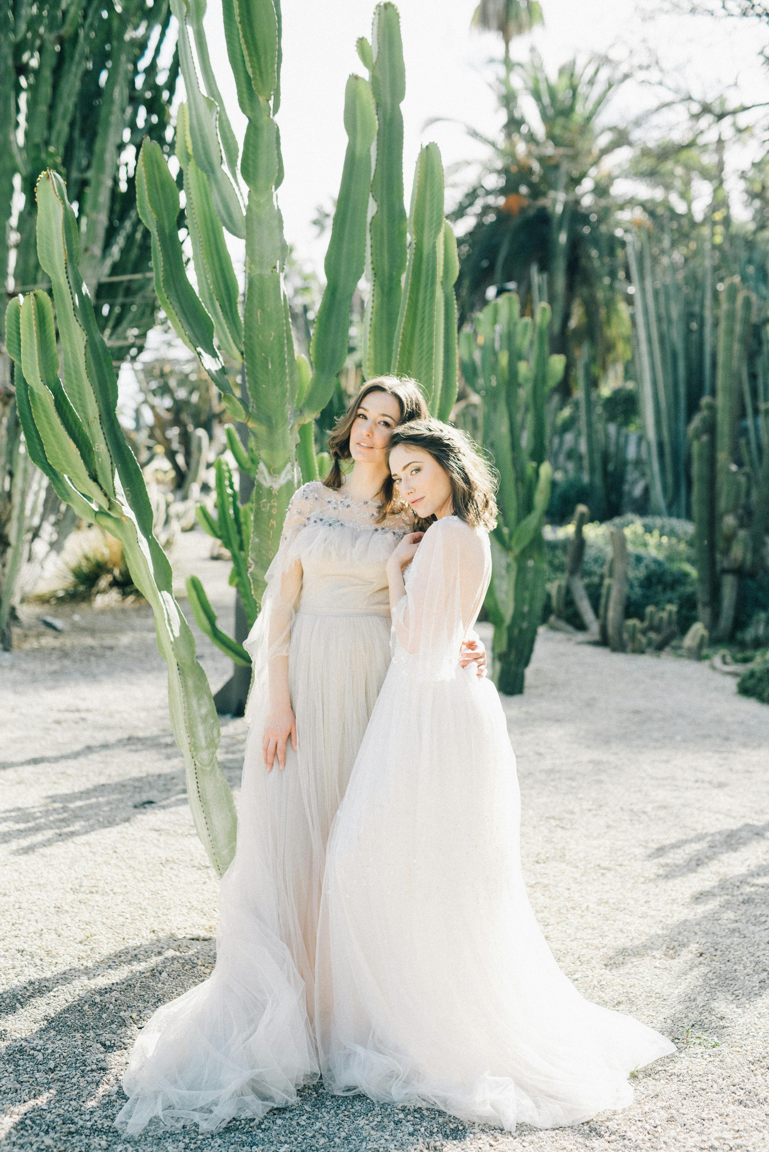 Two women in elegant dresses pose gracefully by towering cacti, showcasing serene beauty.