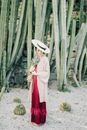 一个戴着草帽、身穿红色连衣裙的女人，手持树叶，站在高高的仙人掌中间