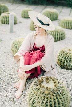 一位戴着草帽、身着红色连衣裙与和服的优雅女子，坐在桶仙人掌中间