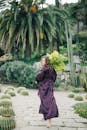 一位身穿紫色缎面长袍的女子，手捧花束，在郁郁葱葱的花园中微笑前行