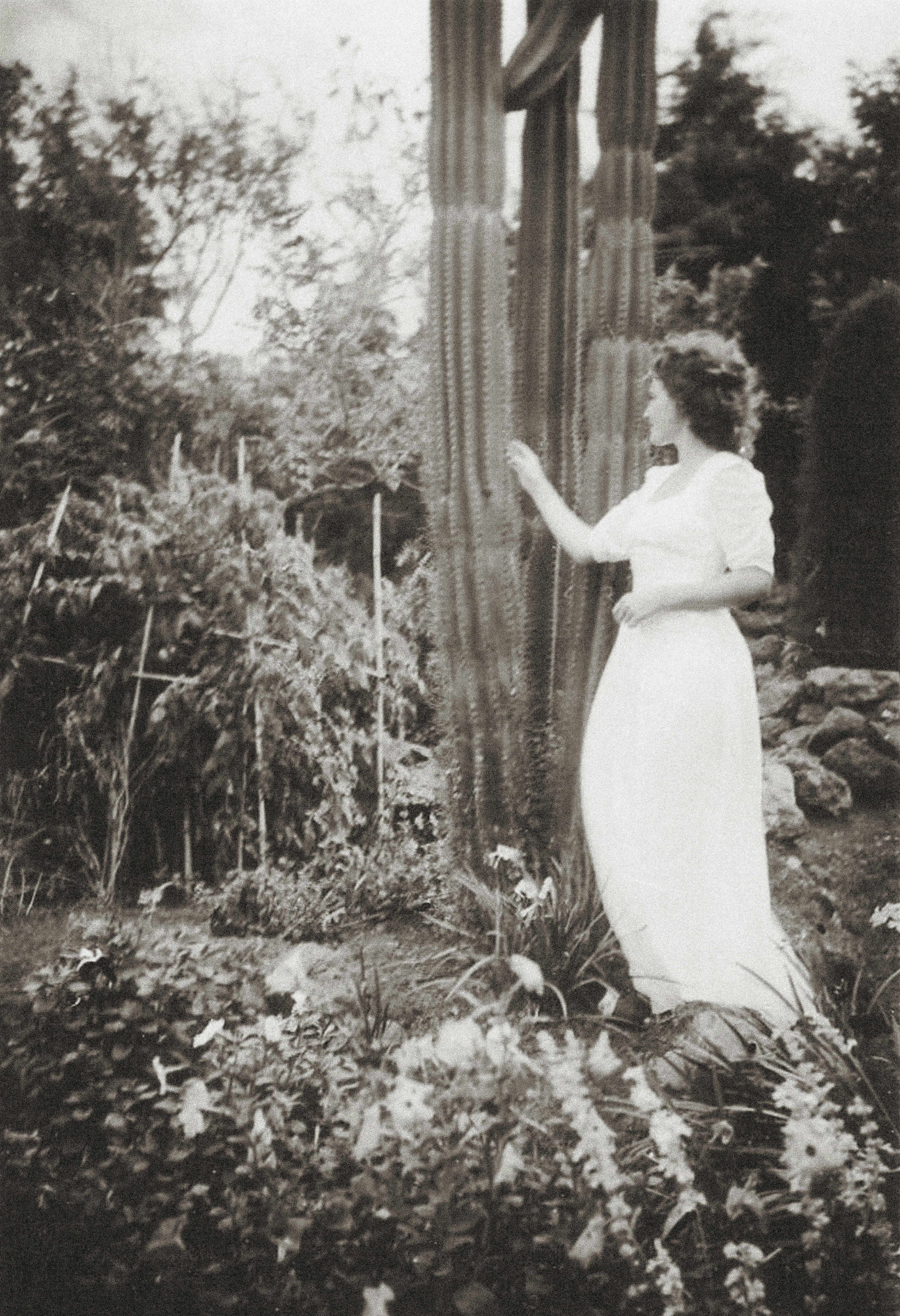 A vintage photo of a woman in a white dress standing beside a large cactus in a garden.
