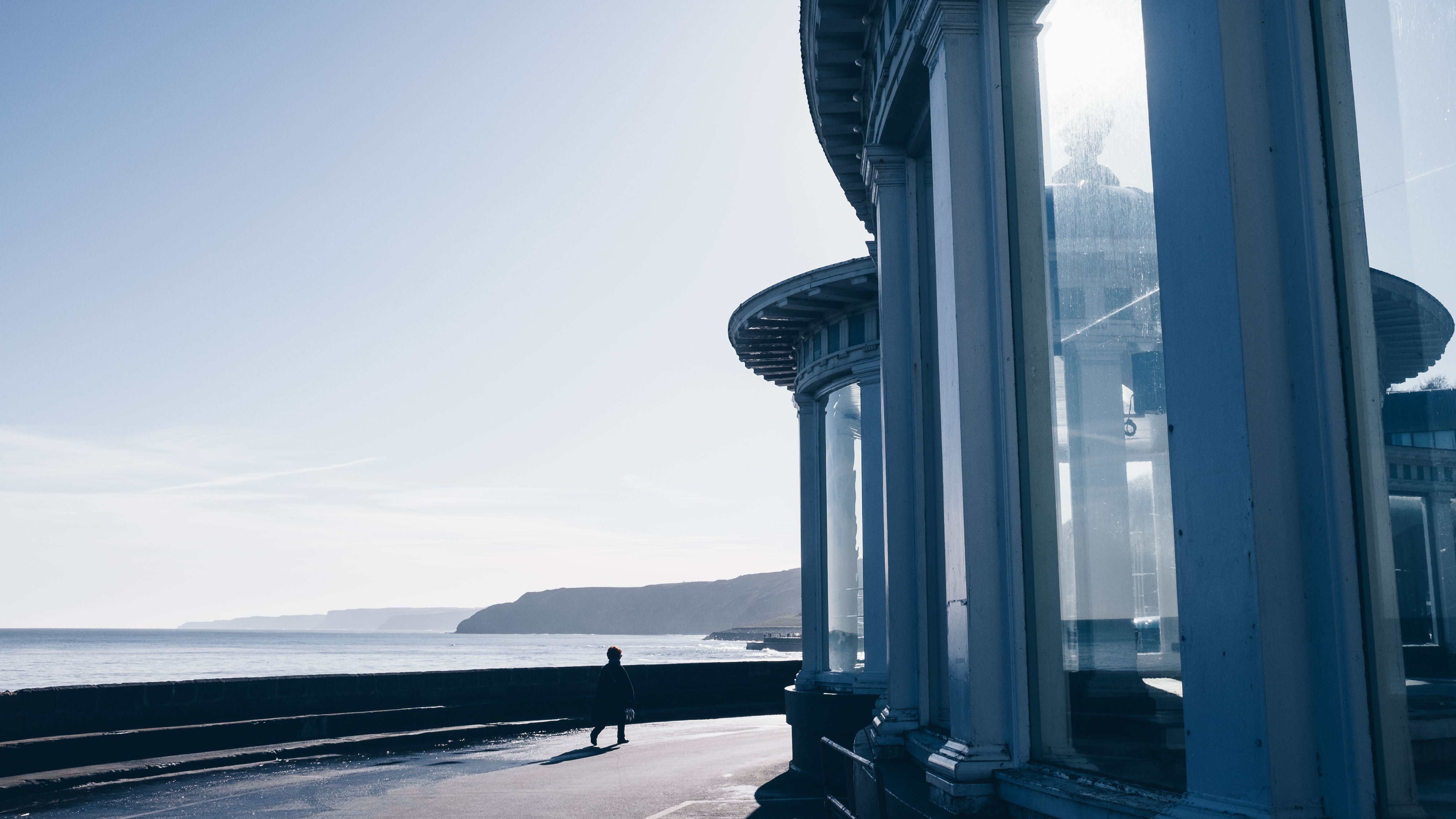 A lone walker passes a curved glass pavilion on Aberystwyth seafront, United Kingdom, beneath bright winter light and coastal hills.