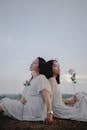 Side view of dreamy adult Asian woman holding hand of young daughter while sitting together back to back in field with roses in hands