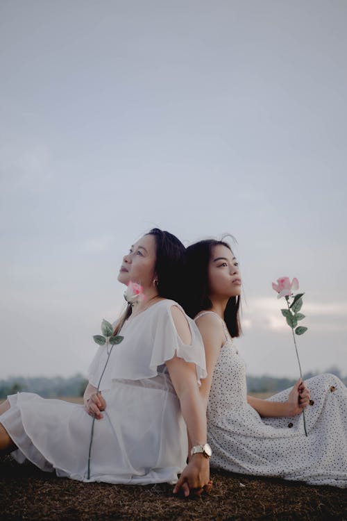 Free Side view of dreamy adult Asian woman holding hand of young daughter while sitting together back to back in field with roses in hands Stock Photo