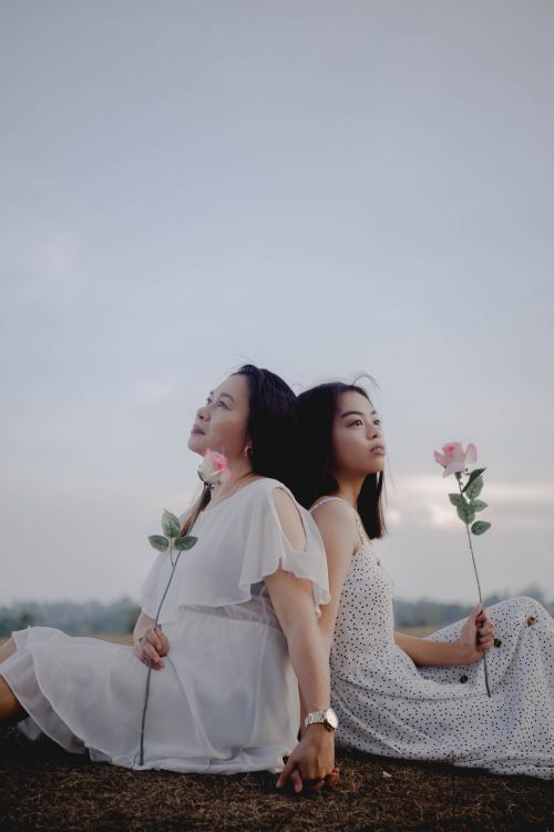 Free Side view of dreamy adult Asian woman holding hand of young daughter while sitting together back to back in field with roses in hands Stock Photo