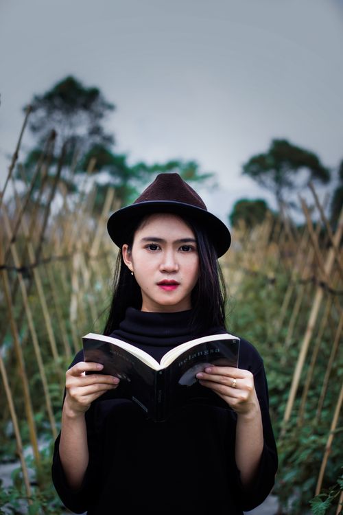 Free Attentive young Asian female in black outfit and hat standing in garden and reading interesting book against cloudy sky Stock Photo