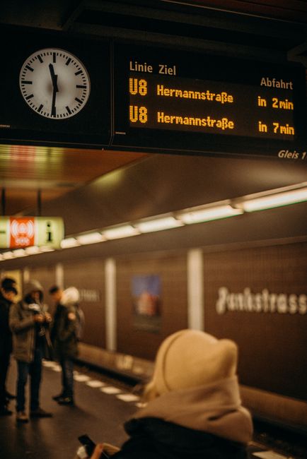 A bustling Berlin subway platform scene with people waiting and schedules visible.