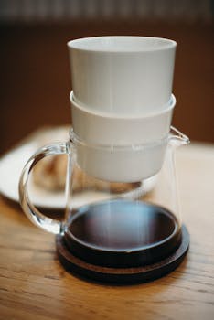 A minimalist image of a clear glass coffee jug with a white ceramic cup set on a wooden table.