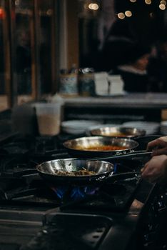 A chef expertly handles frying pans over open flames in a bustling kitchen setting.