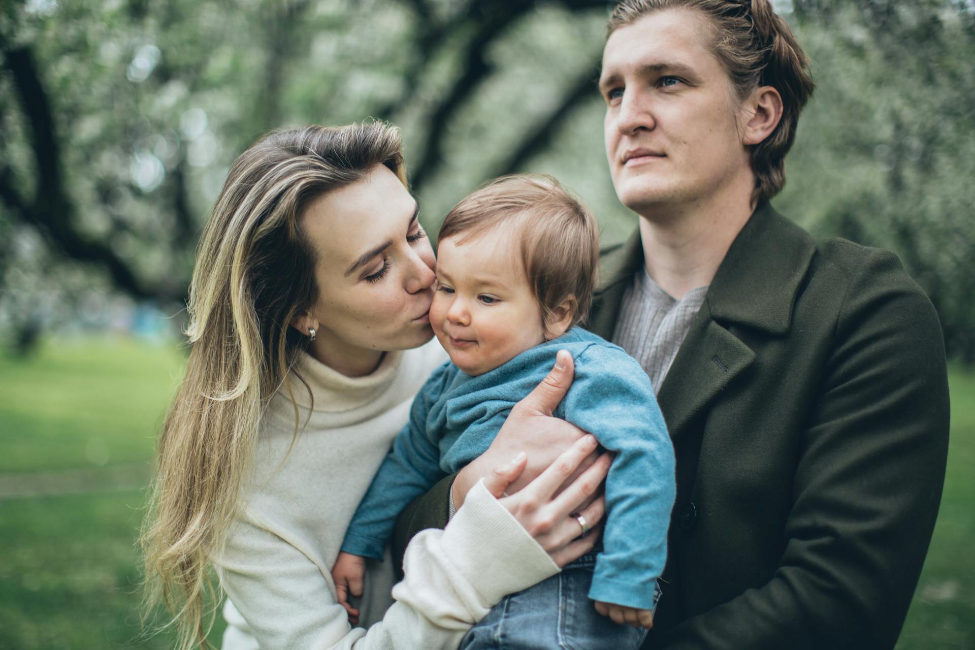 Mother kissing baby with father nearby, family enjoying a day outdoors.