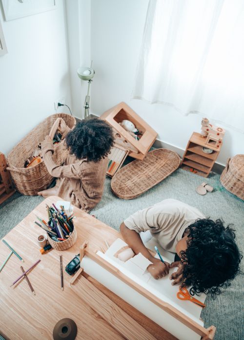 Free Top view of anonymous African American child drawing on paper sheet with pencil while sitting near barefoot sister playing with toys put in wicker basket in flat Stock Photo