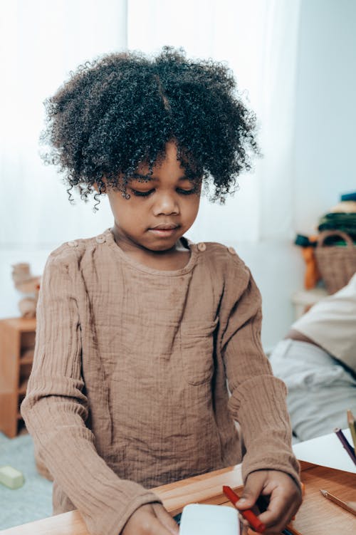 From above of adorable pensive black kid with Afro hairstyle in casual clothes sitting with white paper cube and red felt pen at wooden table at home and looking down