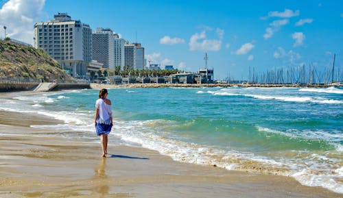 Back view of full length of female tourist in summer outfit strolling on sandy beach along ocean with waves in modern city with contemporary buildings in sunny day