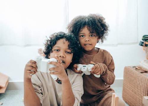 Adorable African American sisters in domestic clothes sitting on floor at home and playing video games with gamepad while spending time together and looking at camera